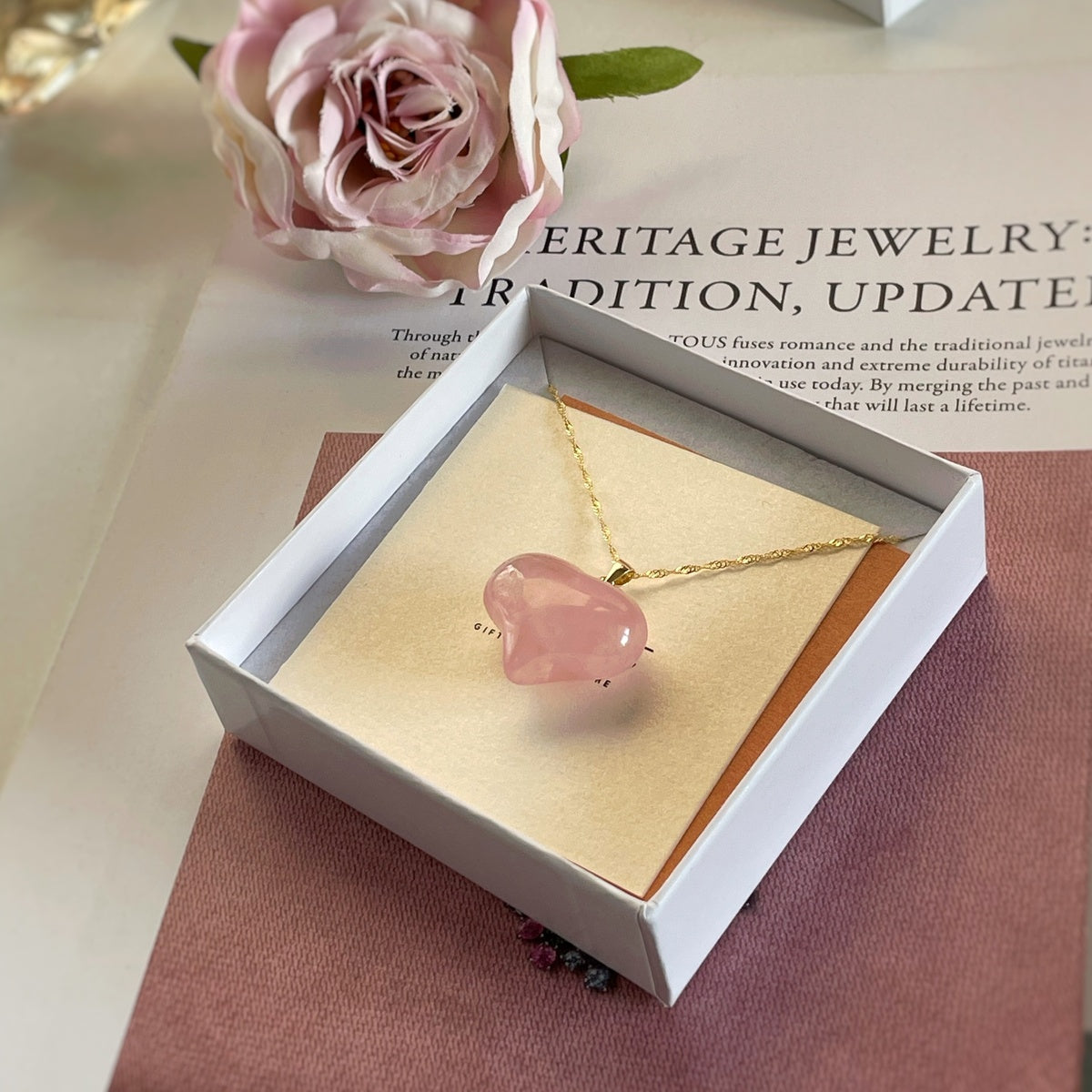 A pink stone pendant necklace on a gold chain, displayed inside an open white jewelry box with a beige insert. The box sits on a pink textured surface with a blurred pink rose and printed paper in the background.