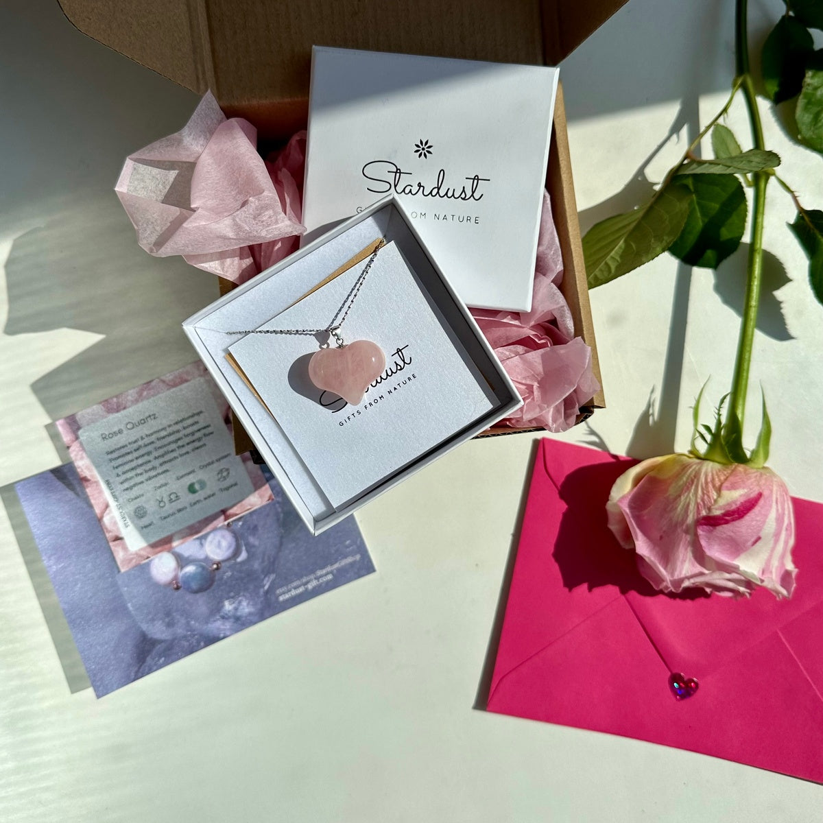 A heart-shaped rose quartz necklace in a white Stardust by Nature packaging box, displayed next to a pink rose and a bright pink envelope on a light surface.